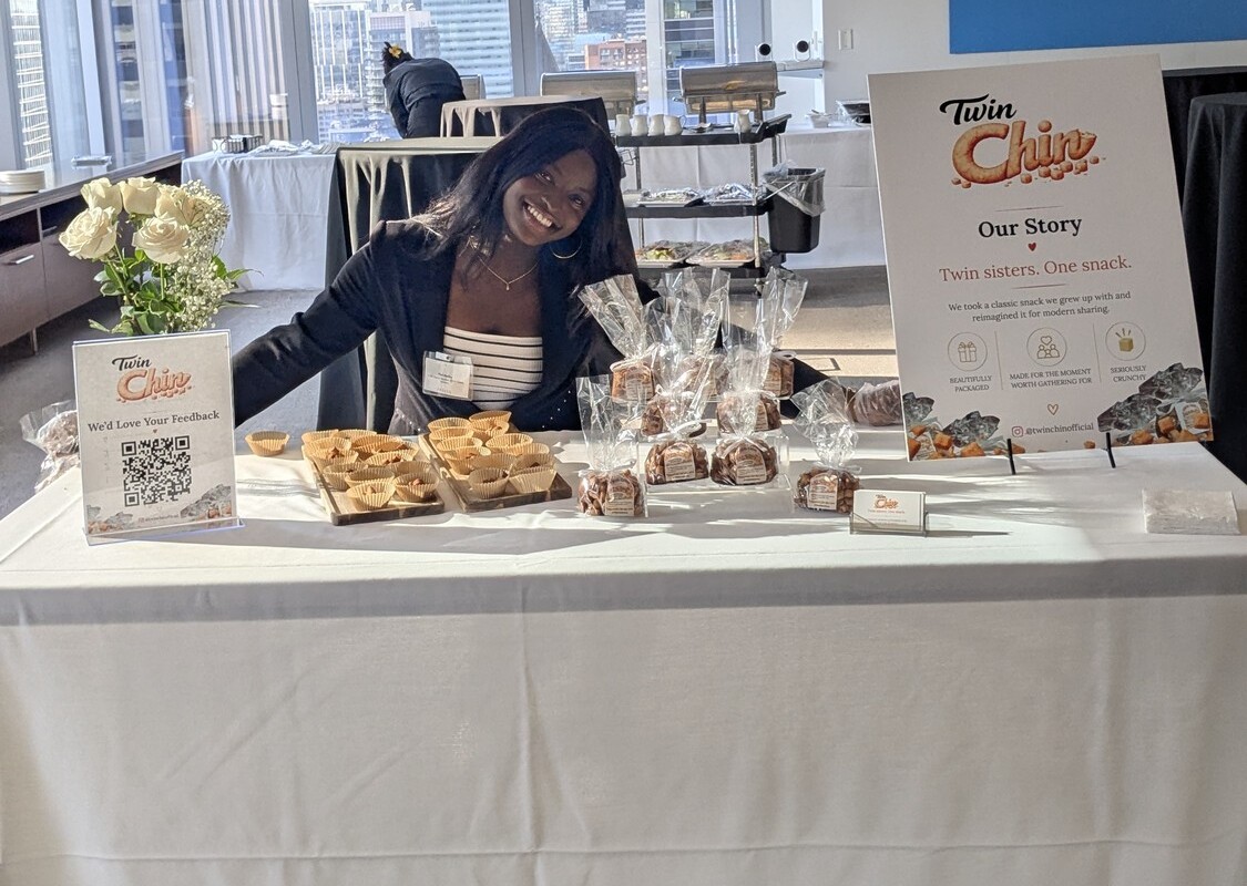 TwinChin founder smiling behind a fully set-up pop-up table with bagged chin chin and signage